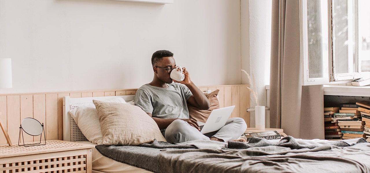 Young man drinking coffee in bed with his laptop